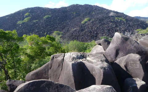 Large dark boulders sit in the foreground with a steep hill covered in black rocks rising behind them, surrounded by patches of green vegetation under a partly cloudy sky.