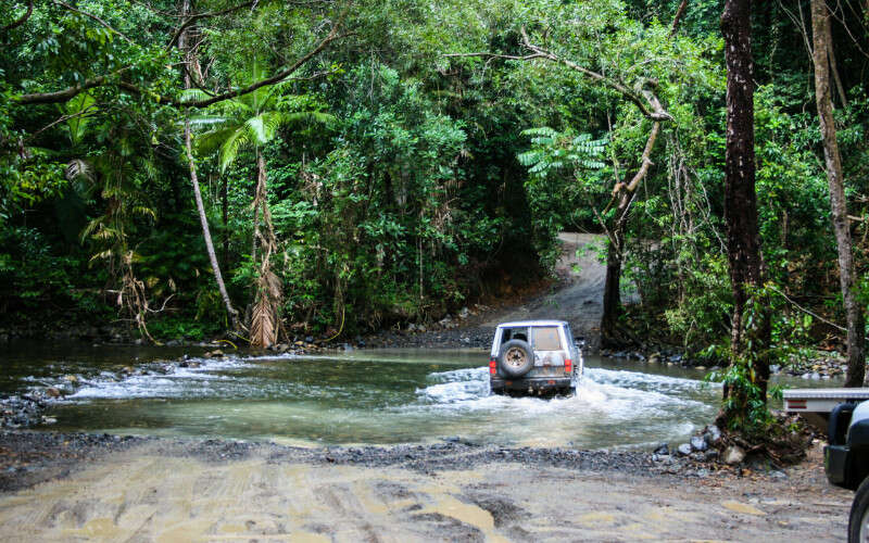 A four-wheel-drive vehicle crosses a shallow river in a dense tropical rainforest, with muddy tracks leading into the water and thick green vegetation surrounding the scene.
