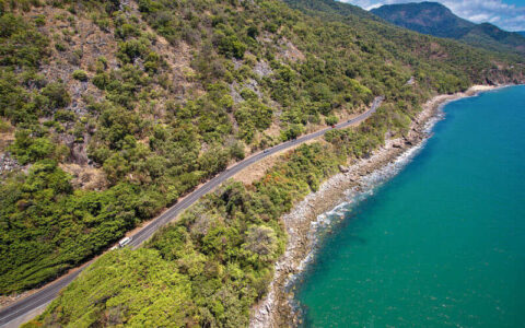 A winding coastal road runs along a steep, forested hillside beside bright blue ocean water, with a vehicle driving along the shoreline route