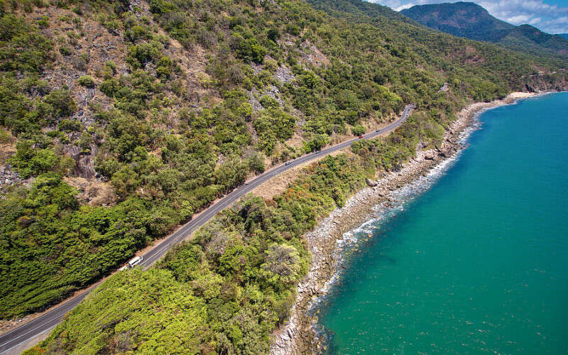 A winding coastal road runs along a steep, forested hillside beside bright blue ocean water, with a vehicle driving along the shoreline route