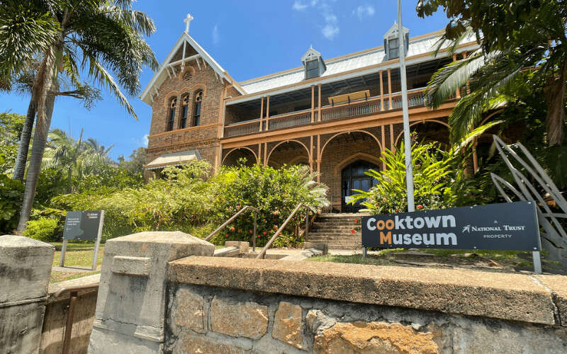 A historic brick building with a veranda and arched details sits behind a sign reading “Cooktown Museum,” surrounded by tropical plants and palm trees under a clear blue sky.