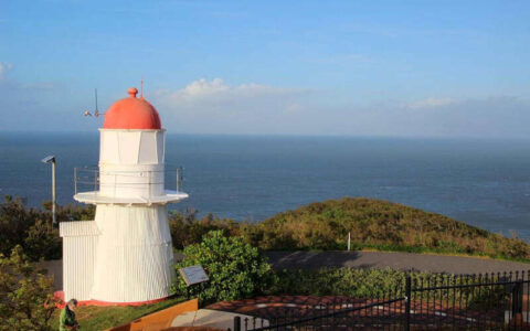 A white lighthouse with a red dome stands on a grassy hill overlooking the ocean, with a coastal road and low vegetation nearby under a clear sky.