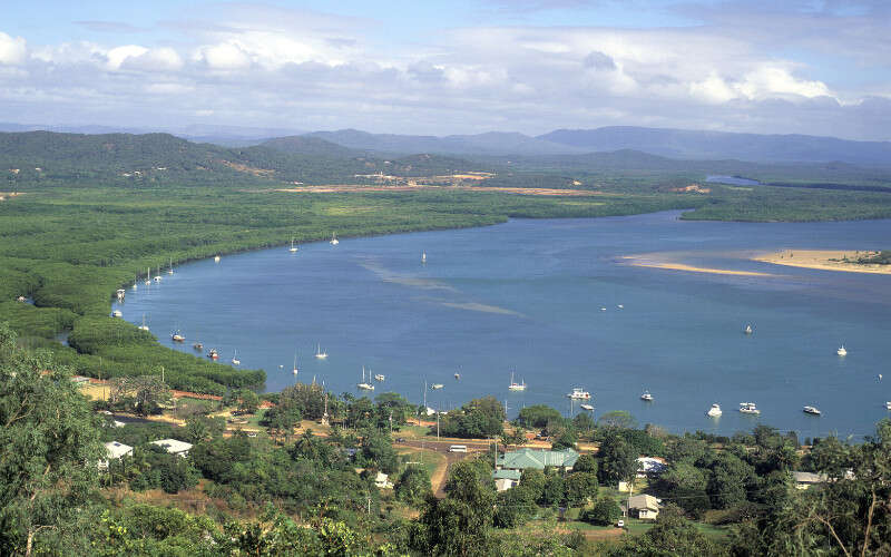 A wide view of a coastal bay with calm blue water dotted with small boats, surrounded by green vegetation, a small town in the foreground, and rolling hills in the distance under a cloudy sky.