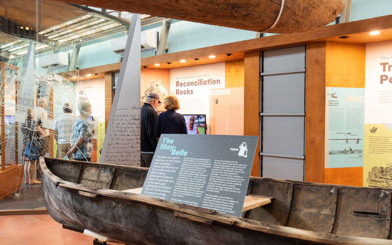 A historic wooden boat is displayed inside a museum gallery, with informational signs and visitors viewing exhibits in the background