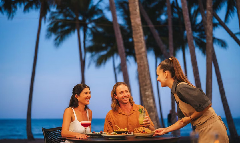 Waitress serving a couple dining at a beachfront restaurant table under palm trees at dusk.