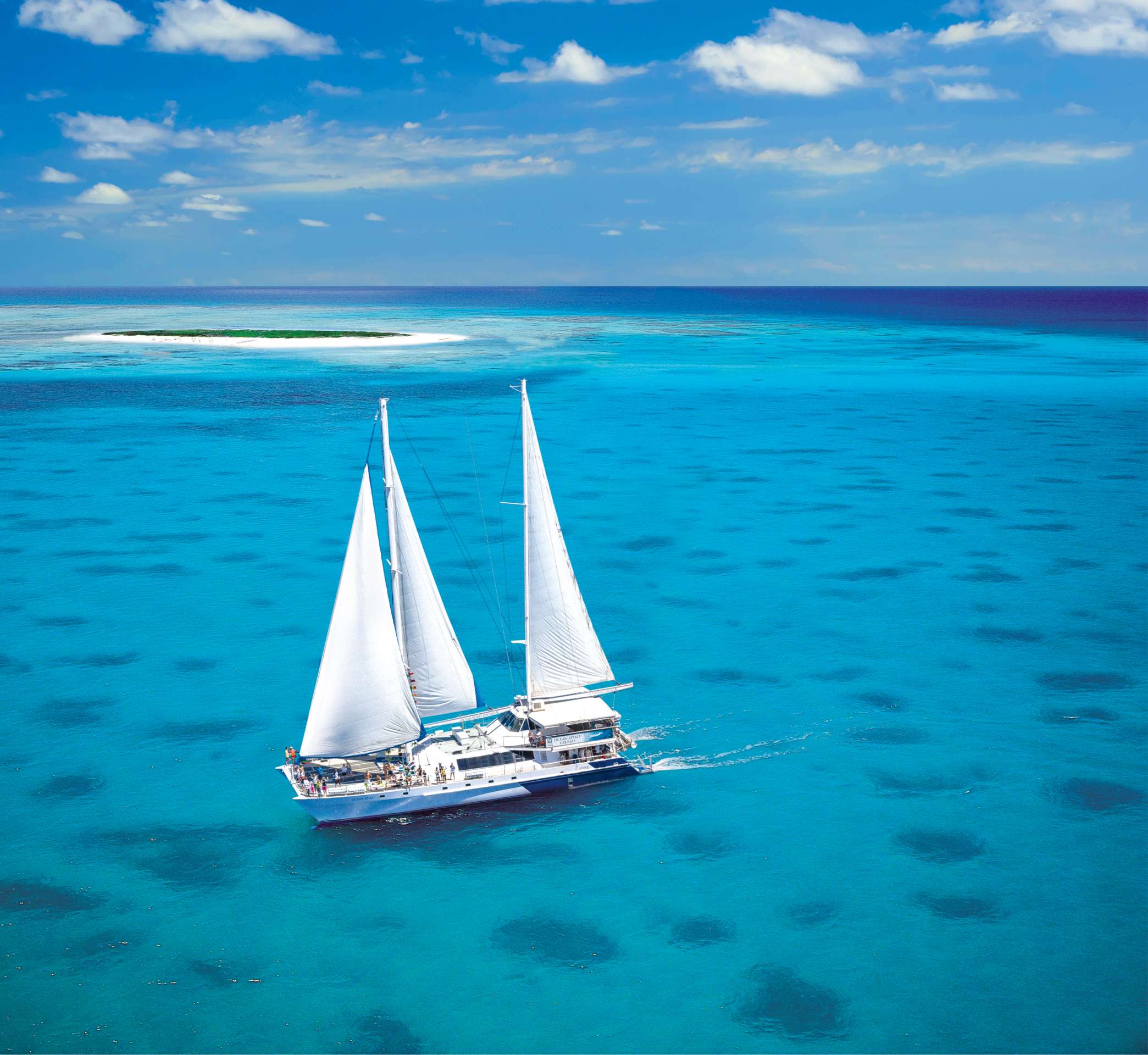 Sailing catamaran with white sails cruising turquoise reef waters near a small sand cay.