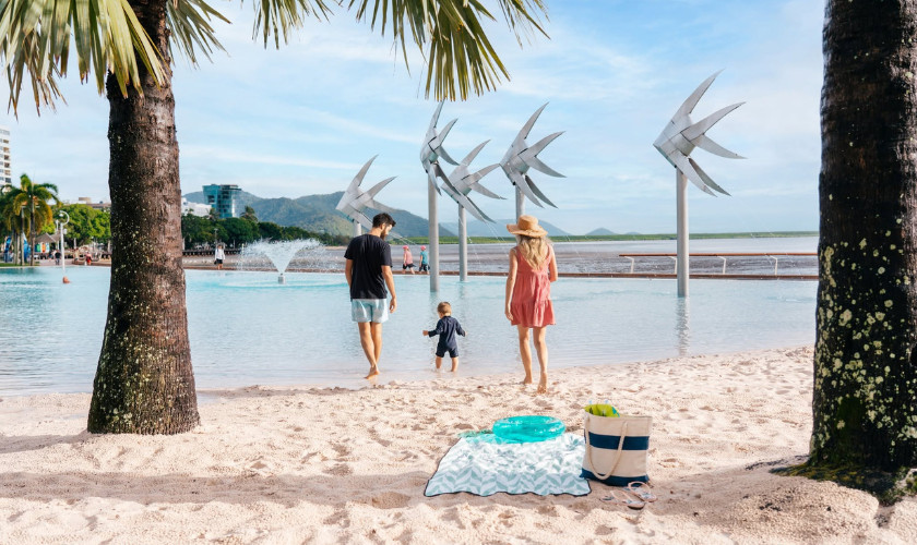 Family walking on the sandy Cairns Esplanade with fish sculptures and the lagoon in the background.