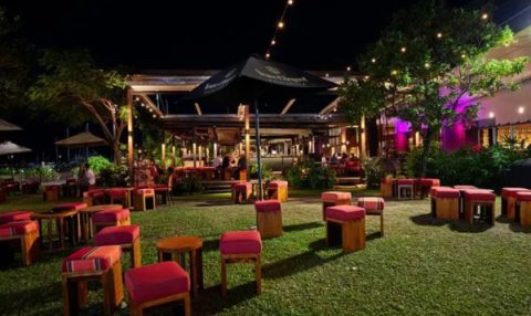 Outdoor restaurant seating area at night with red cushioned stools on grass, lit by string lights and colorful accent lighting.