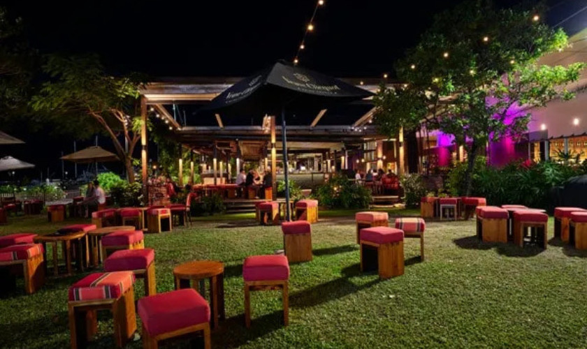 Outdoor restaurant seating area at night with red cushioned stools on grass, lit by string lights and colorful accent lighting.