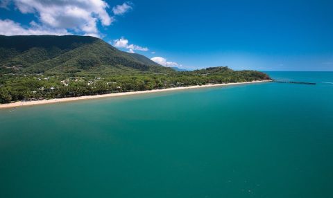 Aerial view of a long sandy beach lined with palm trees, framed by forested hills and turquoise water.