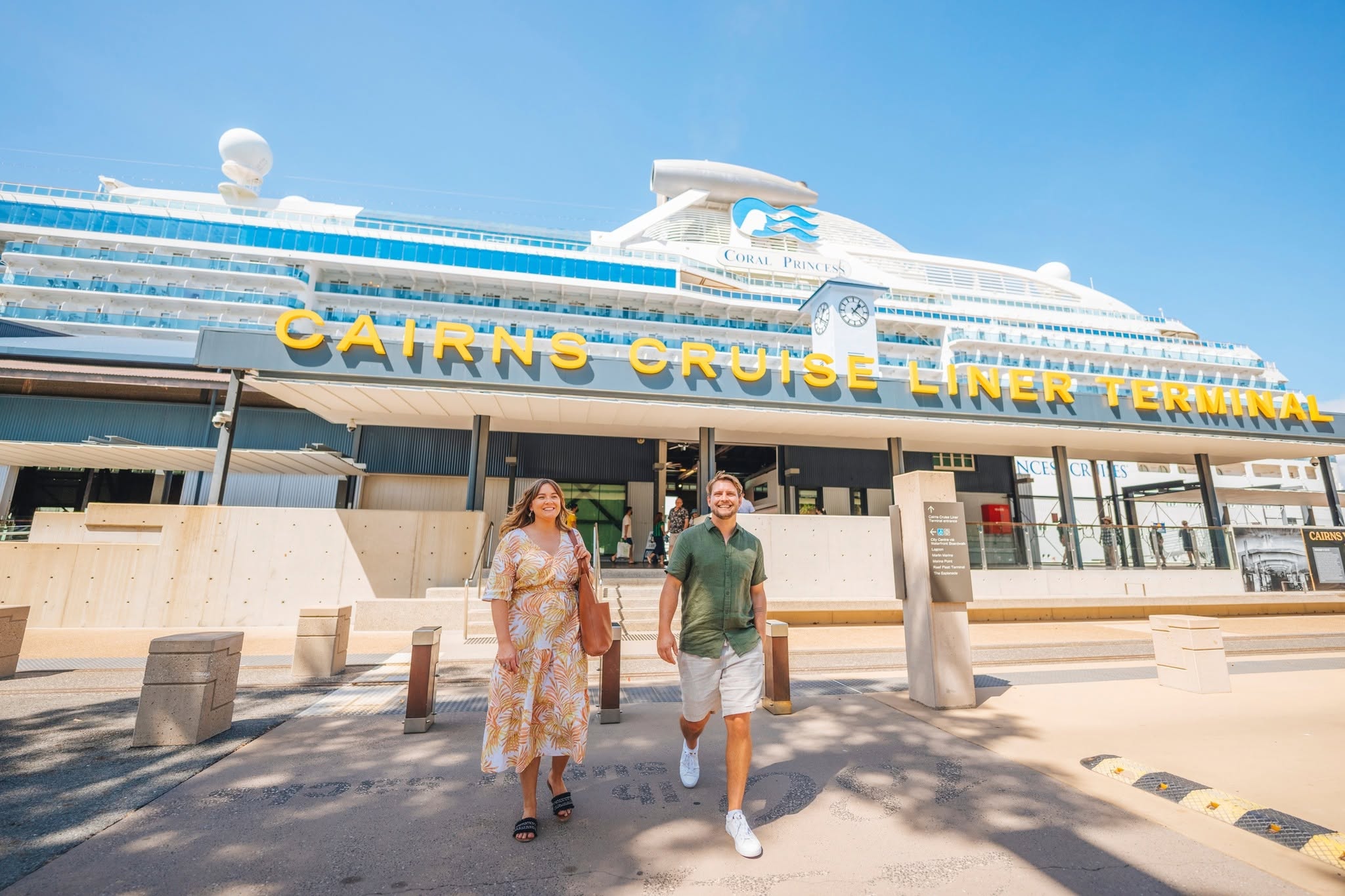 Couple walking away from the Cairns Cruise Liner Terminal building with a large cruise ship docked behind it.