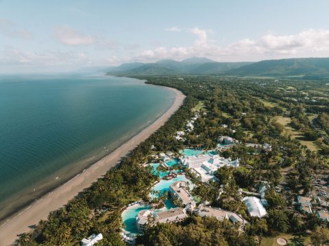 Aerial view of a beachfront resort with pools nestled among coastal rainforest, overlooking a long curved bay.