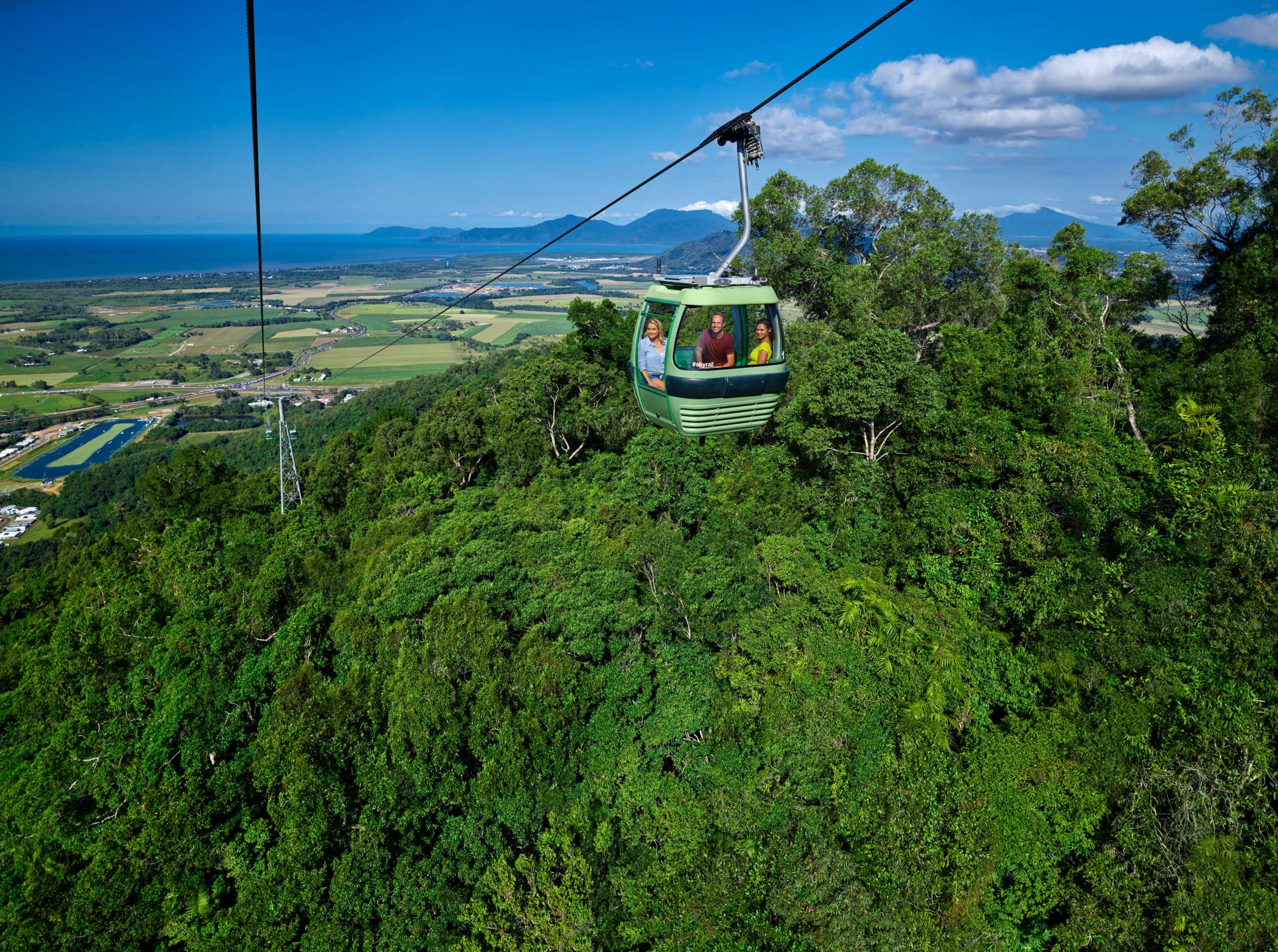 Skyrail cable car gondola over rainforest canopy with coastal farmland and ocean visible in the distance.