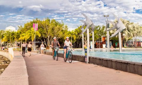 Cyclists and pedestrians on the Cairns Esplanade boardwalk, with large fish sculptures and the swimming lagoon in view.