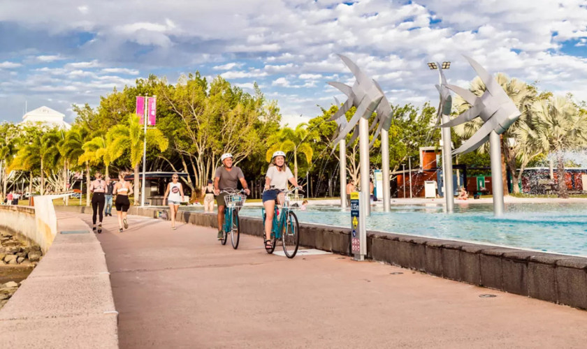 Cyclists and pedestrians on the Cairns Esplanade boardwalk, with large fish sculptures and the swimming lagoon in view.