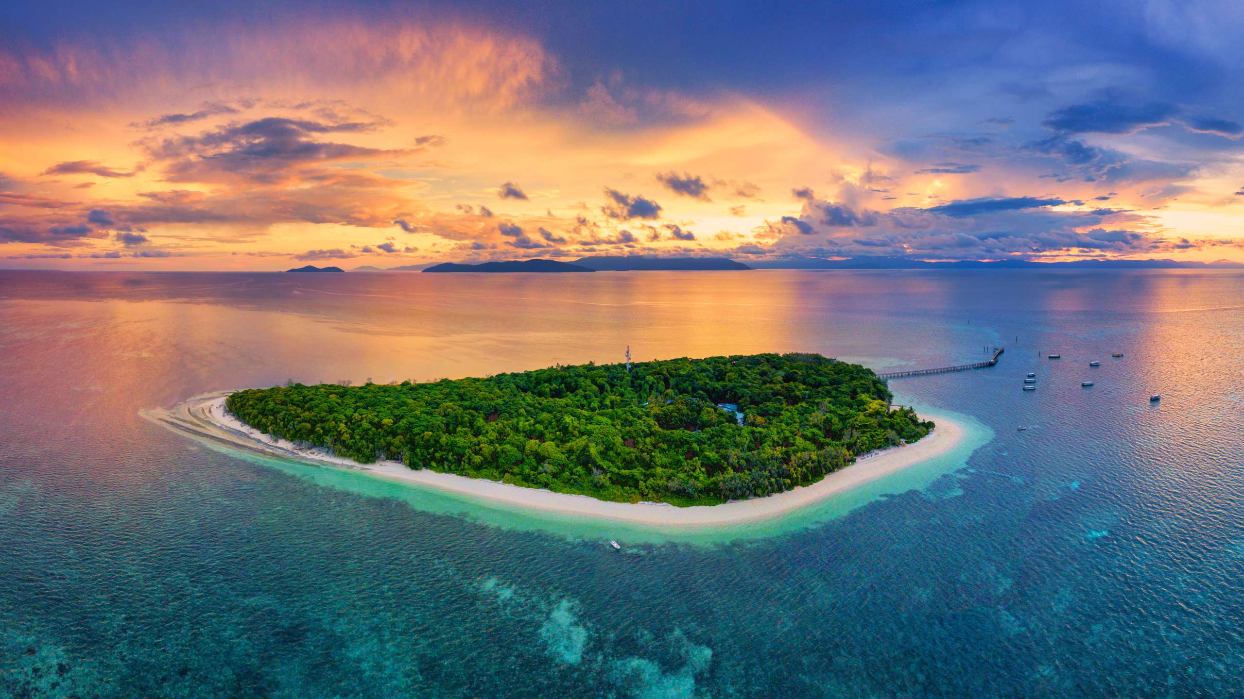 Aerial view of a small tropical island with white sandy beaches and turquoise water at sunset, surrounded by the open ocean.