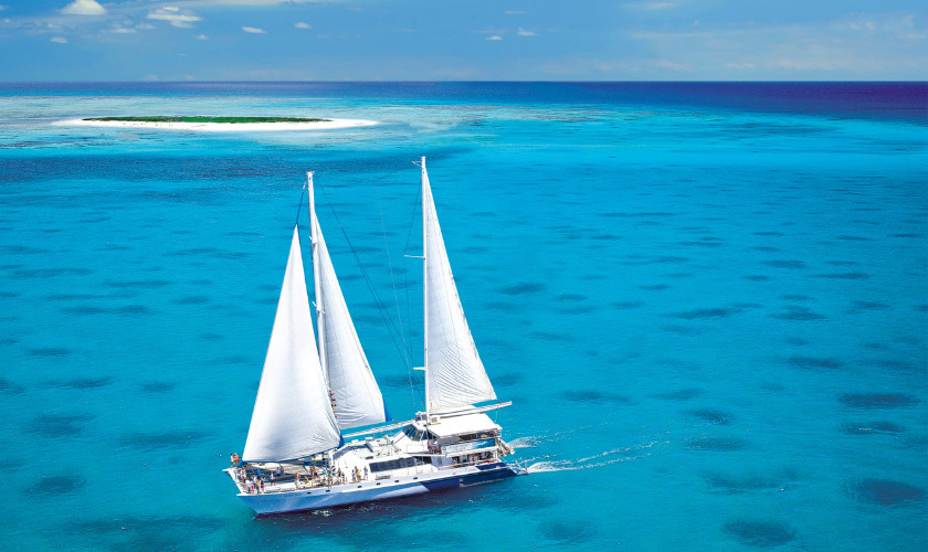 Sailing catamaran with white sails cruising turquoise reef waters near a small sand cay.