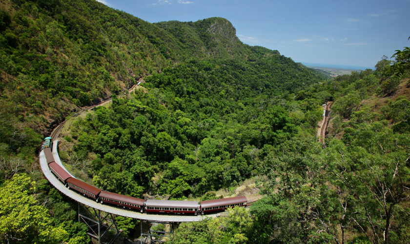 Historic scenic railway train winding along a curved bridge through mountainous rainforest terrain.