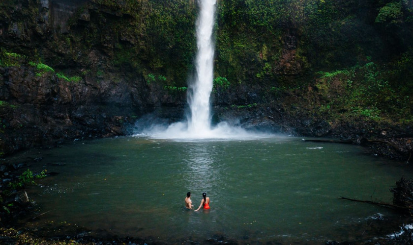 Two people swimming in a plunge pool beneath a tall waterfall surrounded by moss-covered cliffs.
