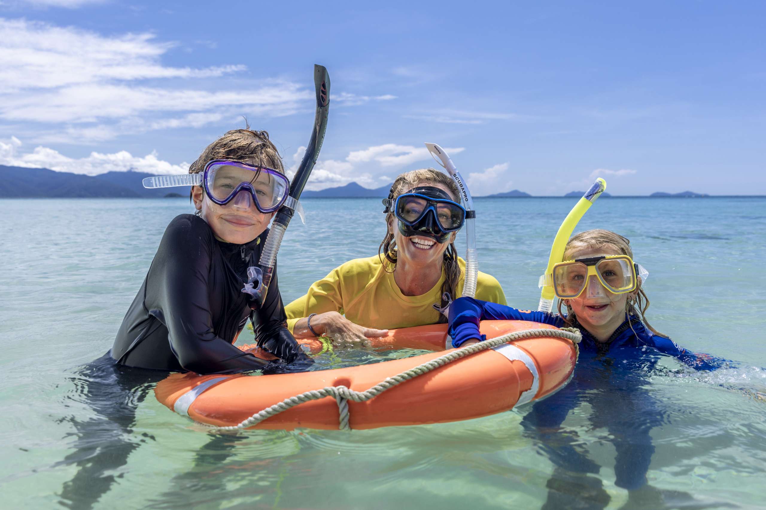 Two children and an adult wearing snorkel masks smile in shallow ocean water, holding onto an orange safety float.
