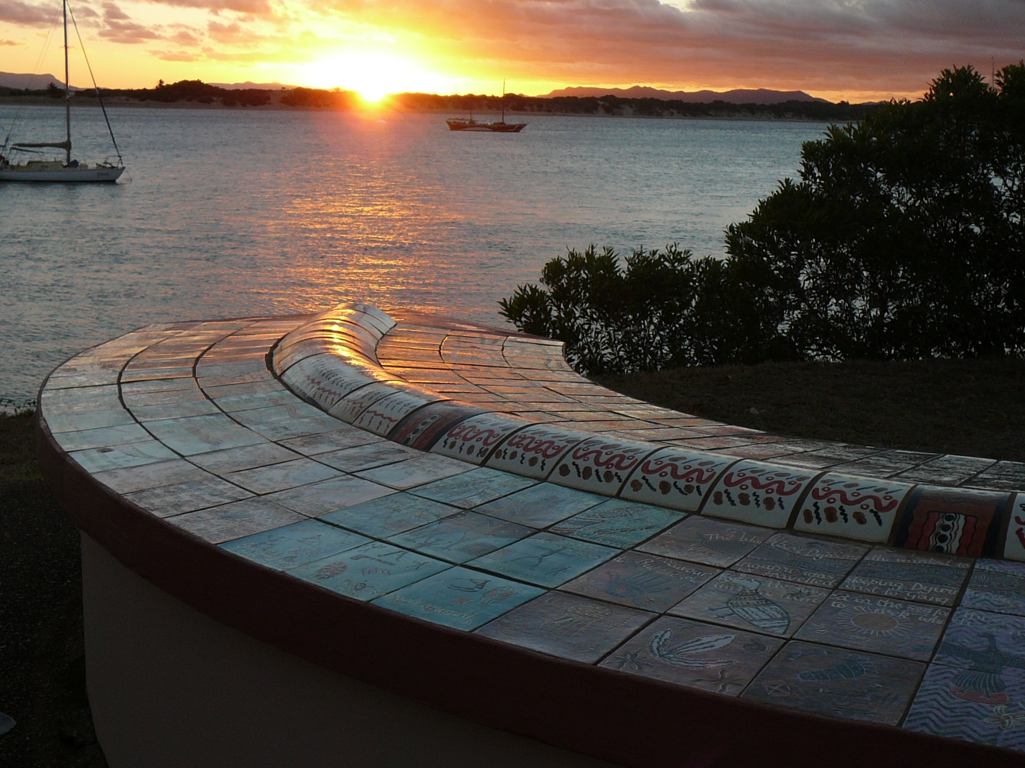 Curved mosaic-tiled bench overlooking a harbor at sunset, with sailboats on the water in the background