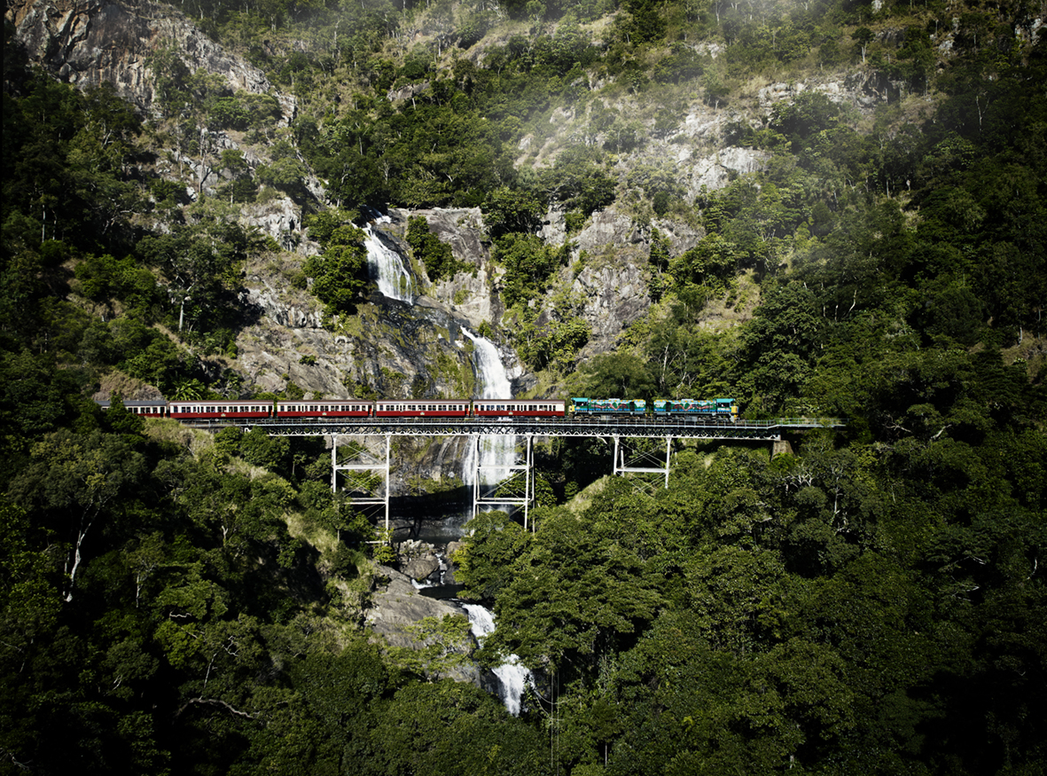 Historic red train crossing a bridge over a waterfall on a lush, forested mountainside.