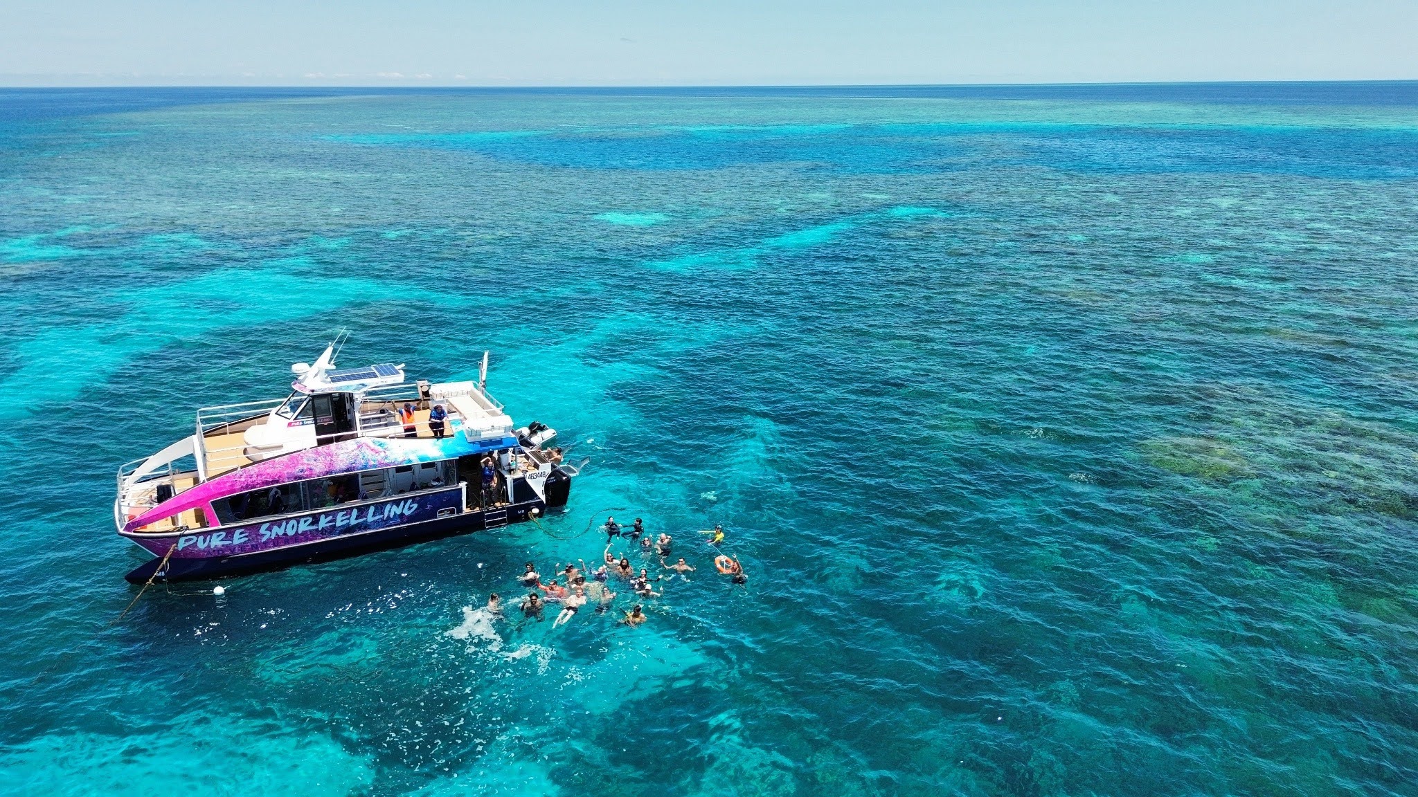 Aerial view of a snorkeling boat with swimmers in the water above vibrant turquoise coral reef.