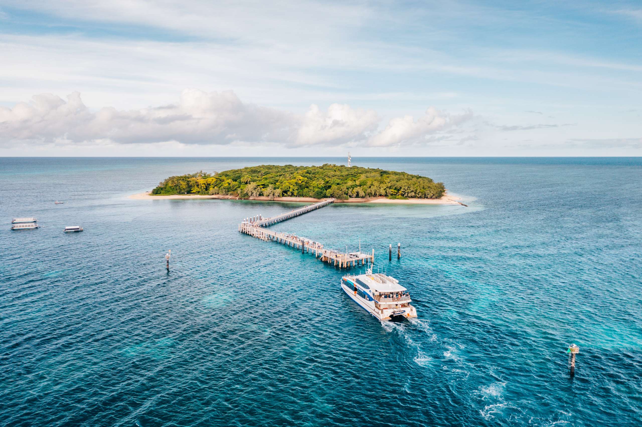 Ferry approaching a small coral cay island via a long jetty, surrounded by clear blue water.