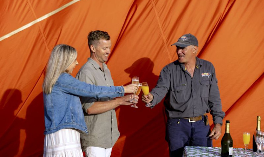 Passengers and pilot toasting with sparkling wine beside a deflated hot air balloon after landing.