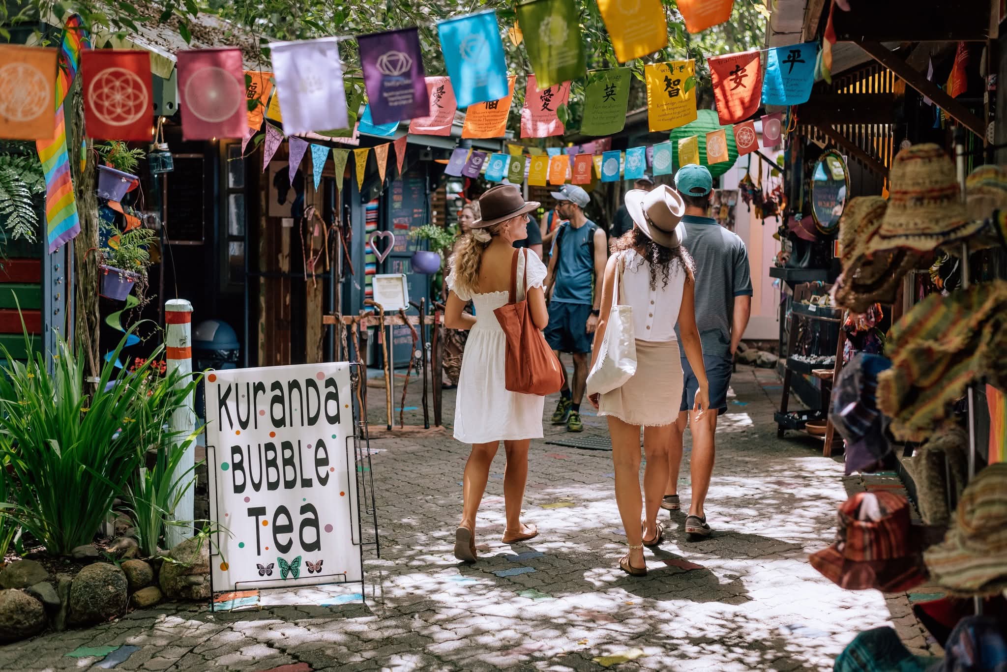 Visitors browsing colorful market stalls in Kuranda Village, decorated with hanging flags and hats.