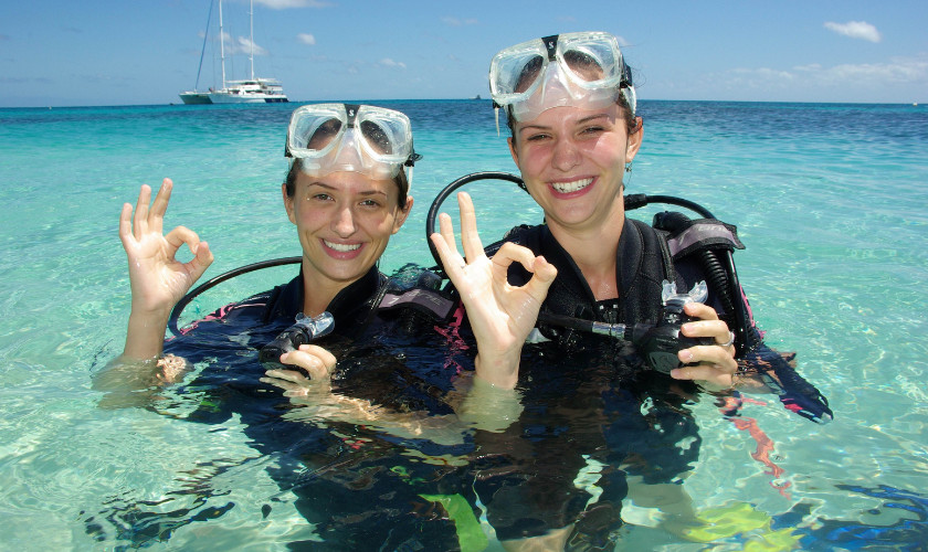 Two divers in scuba gear giving "OK" hand signals while smiling in turquoise water, with a boat in the background.
