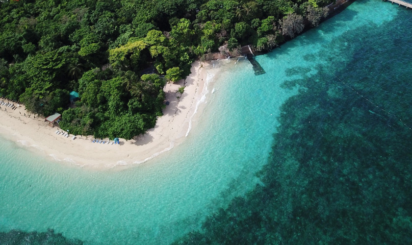 Aerial view of a small forested island with a white sand beach and pier extending into turquoise water.