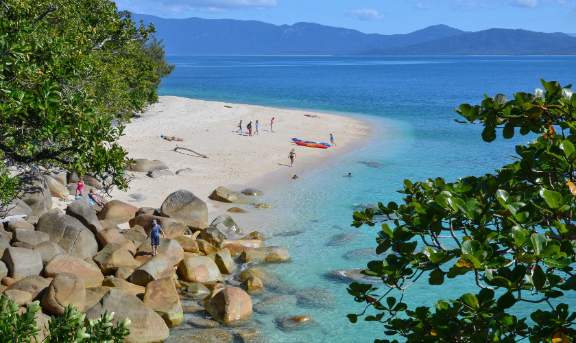 Beachgoers on a rocky, tree-fringed white sand beach with clear turquoise water and mainland mountains in the background.