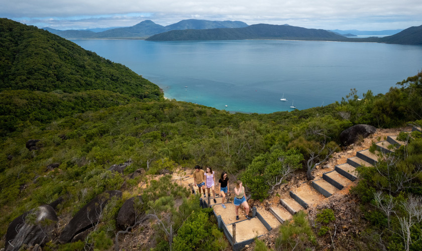 Hikers on a stone stairway trail overlooking a forested island coastline and turquoise bay.