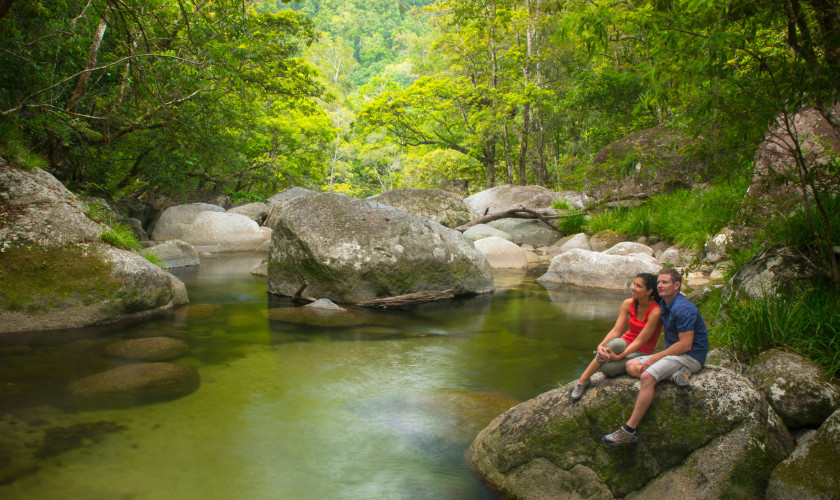 Couple sitting on large boulders beside a tranquil rainforest creek surrounded by dense green foliage.