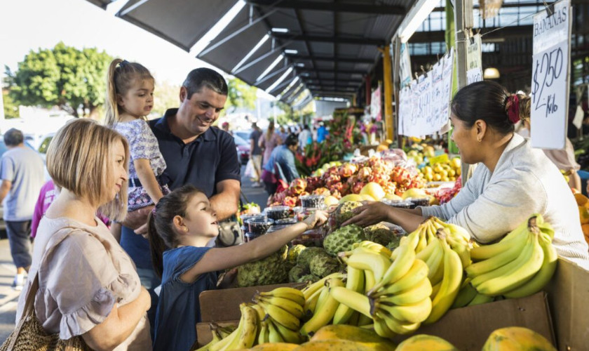 Family buying fresh tropical fruit from a vendor at a busy outdoor market stall.