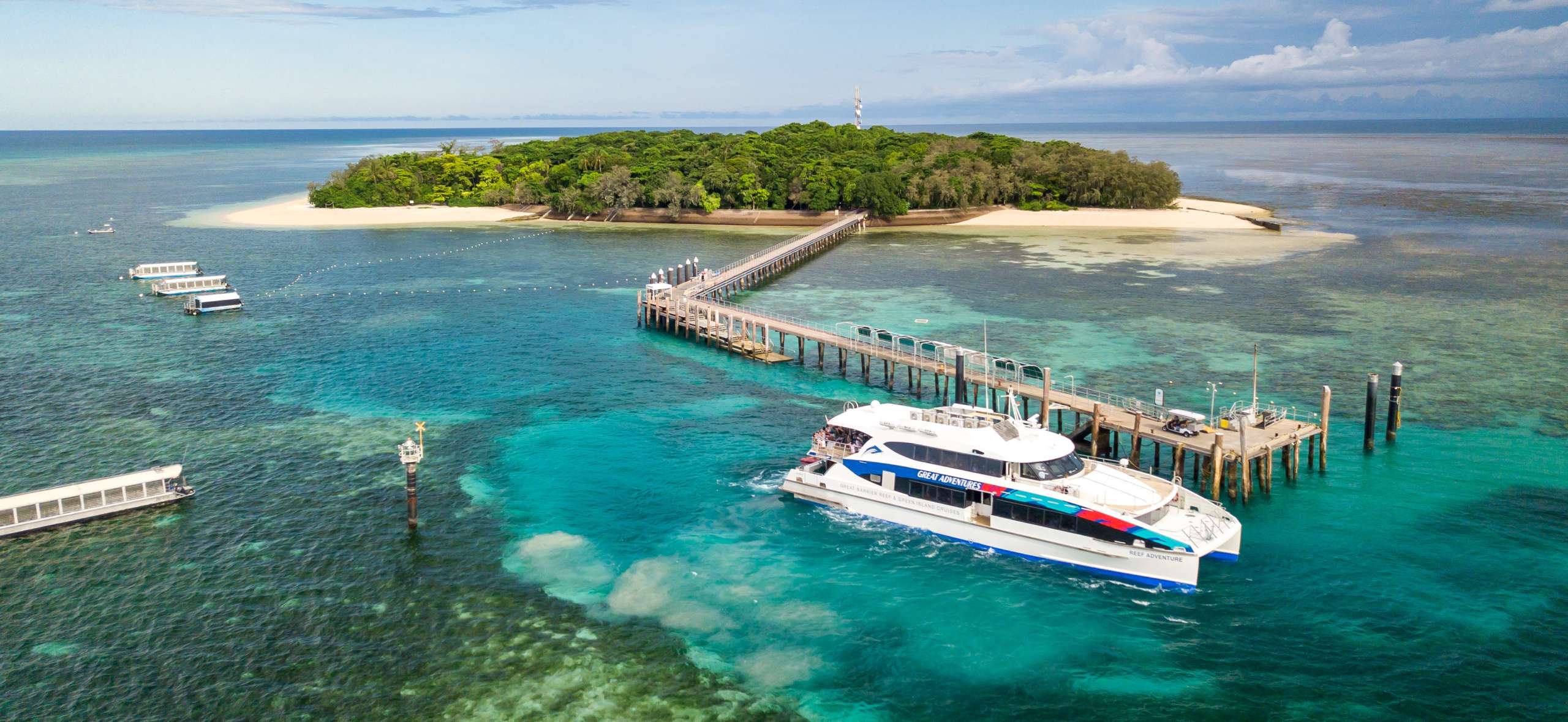 Ferry approaching a small coral cay island via a long jetty, surrounded by clear blue water.