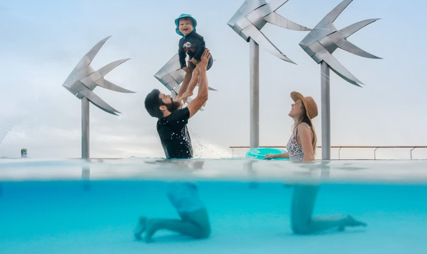Split-view photo of a father lifting a laughing child above the water in the Cairns Esplanade lagoon, with fish sculptures behind them.
